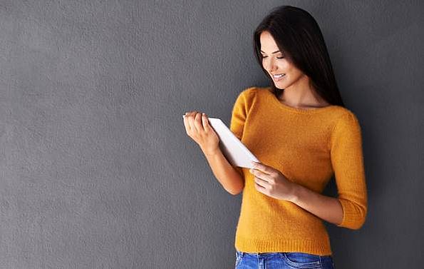Frau mit orangenem Pullover lehnt mit Tablet an grauer Wand