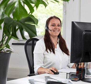 Frau mit Headset sitzt vor dem Bildschirm und lächelt freundlich 