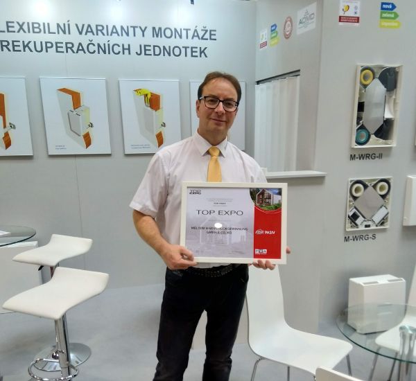 Vertriebsmitarbeiter steht auf Messestand und hält eine Auszeichnung in die Kamera